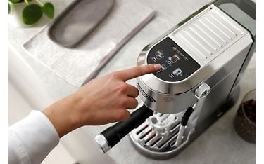 Kuva otettu ylhäältä päin metallin värisestä DeLonghi Dedica Duo -espressokeittimestä. Keitin on keittiötasolla ja henkilön käsi koskettaa koneen digitaalista kosketusnäyttö.