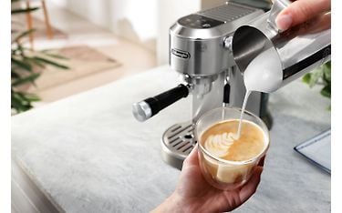 Taustalla näkyy metallin värinen DeLonghi Dedica Duo -espressokeitin. Etualalla henkilön käsissä on lasikupissa kahvia, johon kaataa maitovaahtoa metallisesta kannusta.